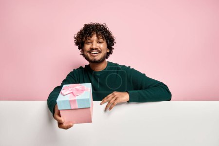 Téléchargez les photos : Homme indien heureux étirant boîte cadeau tout en vous félicitant sur fond rose - en image libre de droit