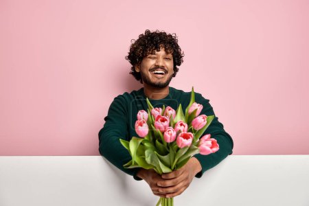 Photo for Handsome Indian man stretching out flower bouquet and winking against pink background - Royalty Free Image