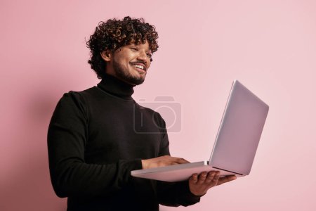 Photo for Handsome Indian man using laptop and smiling against pink background - Royalty Free Image