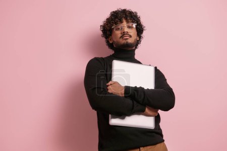 Téléchargez les photos : Confiant jeune homme indien regardant la caméra tout en portant un ordinateur portable sur fond rose - en image libre de droit
