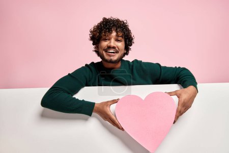 Photo for Happy Indian man holding heart shaped figure and smiling against pink background - Royalty Free Image