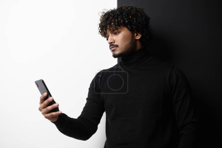 Téléchargez les photos : Beau Indien utilisant un téléphone intelligent tout en se tenant debout sur fond noir et blanc - en image libre de droit