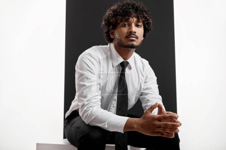 Téléchargez les photos : Homme indien confiant en chemise et cravate regardant la caméra tout en étant assis sur fond noir - en image libre de droit