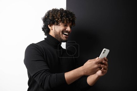 Téléchargez les photos : Homme indien heureux utilisant un téléphone intelligent tout en se tenant debout sur fond noir et blanc - en image libre de droit