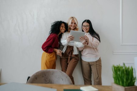 Photo for Three happy mature women looking at digital tablet while leaning on the wall in office together - Royalty Free Image