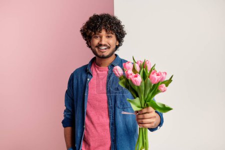 Photo for Happy Indian man stretching out bouquet of tulips against pink and white background - Royalty Free Image