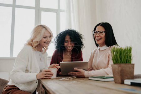 Téléchargez les photos : Trois femmes mûres heureuses utilisant une tablette numérique assis au bureau ensemble - en image libre de droit