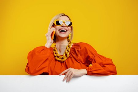 Téléchargez les photos : Jeune femme à la mode dans des lunettes à la mode tenant le téléphone intelligent sur fond jaune - en image libre de droit