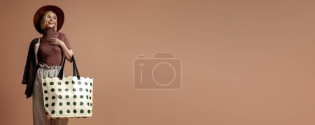 Téléchargez les photos : Jolie jeune femme au chapeau élégant portant un sac à la mode et souriant sur fond brun - en image libre de droit