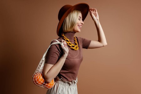 Photo for Joyful young woman in elegant hat carrying mesh bag with oranges against brown background - Royalty Free Image