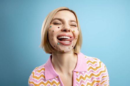 Foto de Mujer joven feliz con diamantes de imitación de colores sobre su cara sobresaliendo lengua contra fondo azul - Imagen libre de derechos