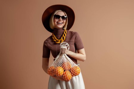 Téléchargez les photos : Joyeuse jeune femme dans un élégant chapeau portant un sac en filet avec des oranges sur fond brun - en image libre de droit