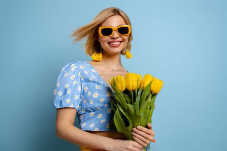 Téléchargez les photos : Joyeux jeune femme dans des lunettes à la mode tenant tas de tulipes jaunes sur fond bleu - en image libre de droit
