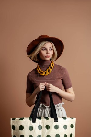 Téléchargez les photos : Jeune femme à la mode dans un chapeau élégant portant sac à la mode sur fond marron - en image libre de droit