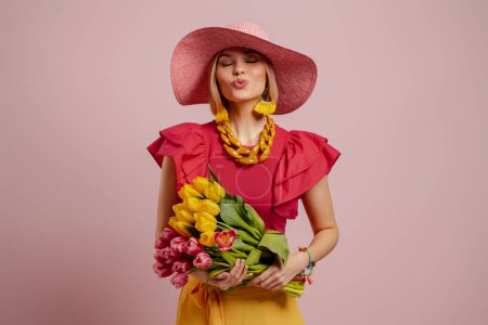 Photo for Elegant young woman blowing a kiss while holding bunch of tulips against pink background - Royalty Free Image