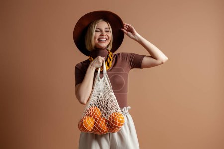 Téléchargez les photos : Joyeux jeune femme dans un élégant chapeau portant un sac en filet avec des oranges sur fond brun - en image libre de droit