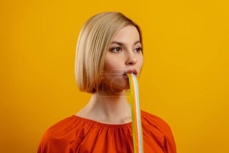 Photo for Beautiful young woman with colorful strips sticking out of mouth standing against yellow background - Royalty Free Image