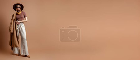 Téléchargez les photos : Pleine longueur de jeune femme à la mode dans un chapeau élégant debout sur fond brun - en image libre de droit