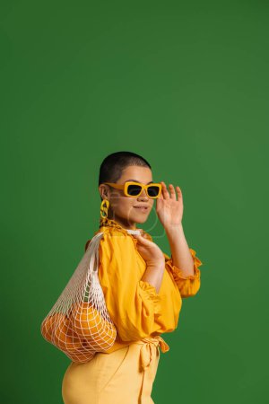 Téléchargez les photos : Jolie jeune femme aux cheveux courts portant un sac en filet avec des oranges sur fond vert - en image libre de droit