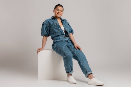 Photo for Happy young short hair woman in denim clothes sitting against grey background - Royalty Free Image