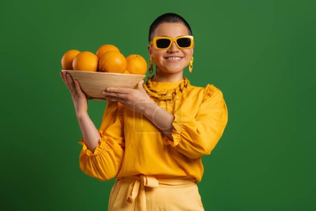Téléchargez les photos : Jeune femme aux cheveux courts à la mode portant une assiette avec des oranges sur fond vert - en image libre de droit