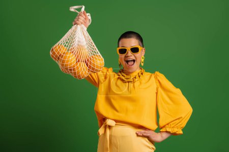 Téléchargez les photos : Jeune femme aux cheveux courts excitée montrant son sac en maille avec des oranges sur fond vert - en image libre de droit
