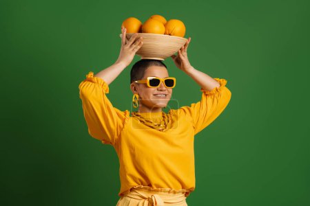 Téléchargez les photos : Joyeux jeune femme cheveux courts portant plaque avec des oranges sur la tête sur fond vert - en image libre de droit