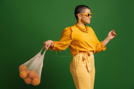 Téléchargez les photos : Jeune femme aux cheveux courts ludique portant un sac en filet avec des oranges sur fond vert - en image libre de droit