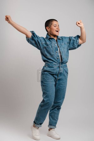 Téléchargez les photos : Pleine longueur de jeune femme excitée aux cheveux courts en vêtements en denim dansant sur fond gris - en image libre de droit