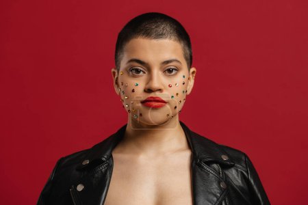 Photo for Beautiful shaved head woman with shiny crystals over her face standing against red background - Royalty Free Image