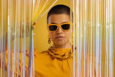Téléchargez les photos : Femme à la mode dans des lunettes à la mode regardant à travers un rideau coloré sur fond jaune - en image libre de droit