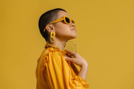 Téléchargez les photos : Jeune femme à la mode aux cheveux courts dans des lunettes à la mode debout sur fond jaune - en image libre de droit