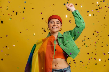 Téléchargez les photos : Femme heureuse portant drapeau arc-en-ciel et geste tandis que confettis volant sur fond jaune - en image libre de droit