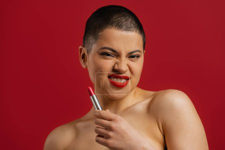 Téléchargez les photos : Jeune femme aux cheveux courts ludique faisant un visage tout en tenant du rouge à lèvres sur fond rouge - en image libre de droit