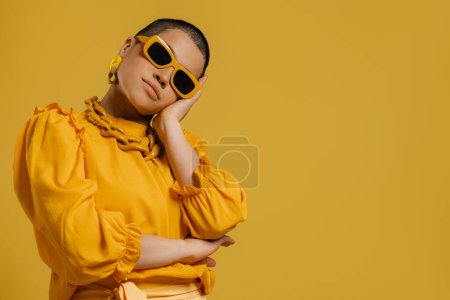 Téléchargez les photos : Élégante jeune femme aux cheveux courts dans des lunettes à la mode debout sur fond jaune - en image libre de droit