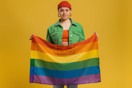 Téléchargez les photos : Jeune femme confiante portant le drapeau arc-en-ciel debout sur fond jaune - en image libre de droit