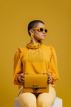 Téléchargez les photos : Femme à la mode dans des lunettes élégantes portant sac à la mode tout en étant assis sur fond jaune - en image libre de droit