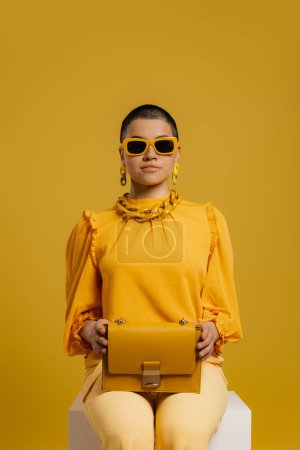 Téléchargez les photos : Femme élégante dans des lunettes à la mode portant sac à la mode tout en étant assis sur fond jaune - en image libre de droit