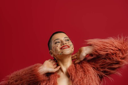 Foto de Feliz joven mujer de pelo corto con cristales brillantes sobre su cara de pie sobre el fondo rojo - Imagen libre de derechos