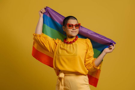Téléchargez les photos : Confiant jeune femme aux cheveux courts tenant drapeau arc-en-ciel sur fond jaune - en image libre de droit