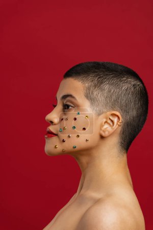 Foto de Vista lateral de la joven cabeza afeitada mujer con cristales brillantes sobre su cara de pie sobre fondo rojo - Imagen libre de derechos