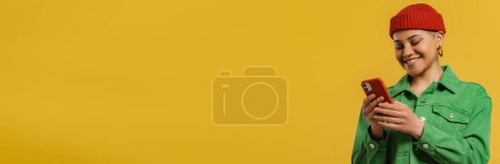 Téléchargez les photos : Jeune femme à la mode utilisant un téléphone intelligent et souriant tout en se tenant debout sur fond jaune - en image libre de droit