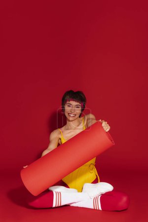 Téléchargez les photos : Joyeuse jeune femme en forme dans un style rétro sportswear tenant tapis d'exercice sur fond rouge - en image libre de droit