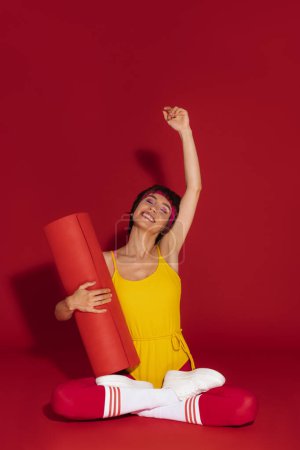 Téléchargez les photos : Happy fit femme en tenue de sport rétro tenant tapis d'exercice et geste sur fond rouge - en image libre de droit