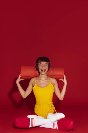 Téléchargez les photos : Heureuse jeune femme en forme dans un style rétro sportswear portant tapis d'exercice sur fond rouge - en image libre de droit
