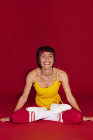 Téléchargez les photos : Heureuse jeune femme en forme dans un style rétro vêtements de sport assis en position lotus sur fond rouge - en image libre de droit