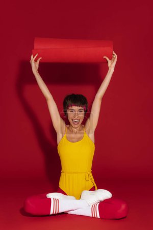 Foto de Mujer joven en forma excitada en ropa deportiva de estilo retro que lleva alfombra de ejercicio contra el fondo rojo - Imagen libre de derechos