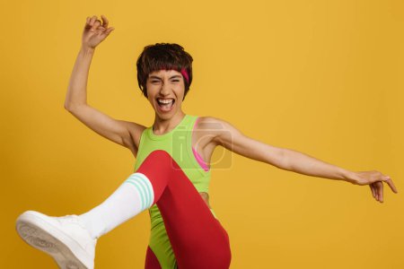 Téléchargez les photos : Femme en forme excitée dans des vêtements de sport de style rétro dansant sur fond jaune - en image libre de droit