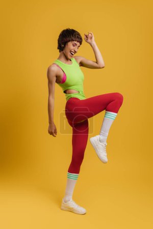 Téléchargez les photos : Pleine longueur de femme en forme excitée dans des vêtements de sport de style rétro exerçant sur fond jaune - en image libre de droit