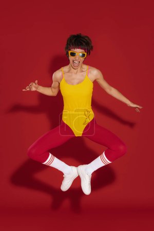 Téléchargez les photos : Heureuse jeune femme dans un style rétro sportswear sautant sur fond rouge - en image libre de droit